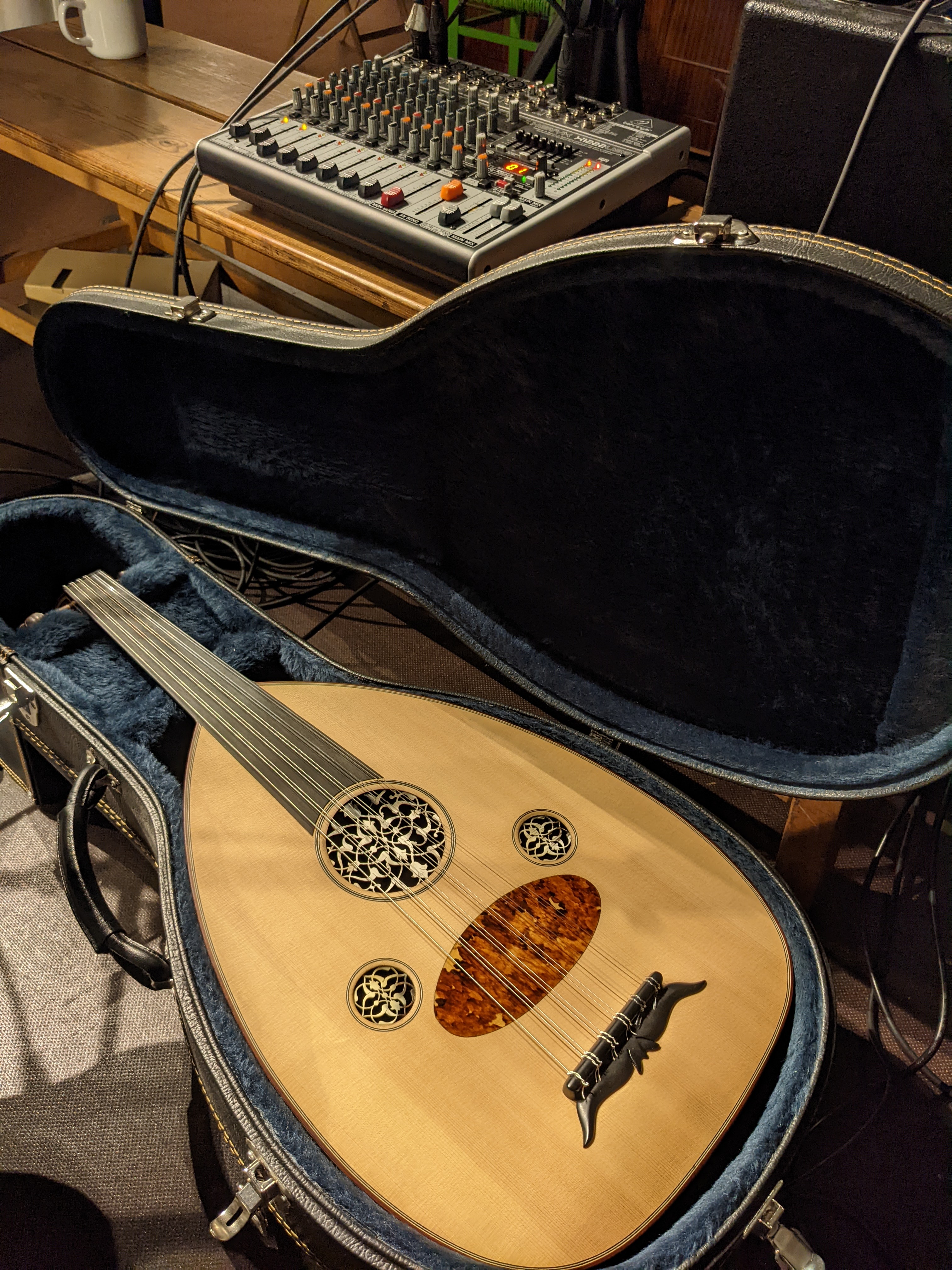 Zu sehen ist eine Mandoline im liegenden aufgeklappten Instrumentenkoffer. Im Hintergrund auf einem Tisch steht ein Mischpult.