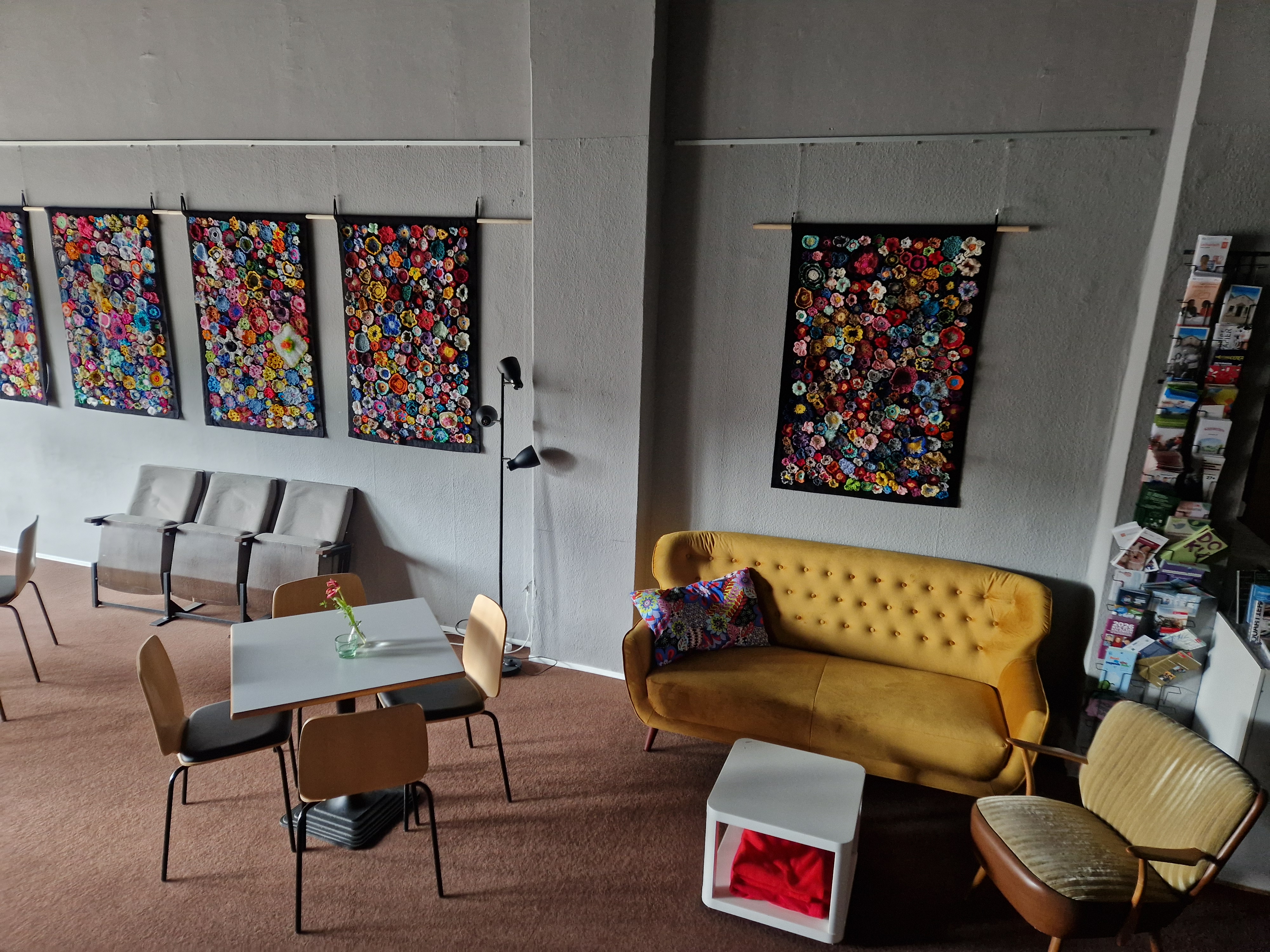 Das NUNATAK von innen: ein gelbes Sofa, Tisch und St&uuml;hle, Sessel. An grau gestrichenen Wand h&auml;ngen Teppiche aus geh&auml;kelten Bl&uuml;ten.